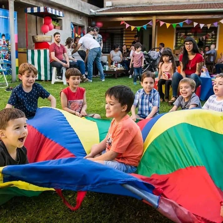 Niños jugando con un colorido paracaídas en una fiesta al aire libre. Otras personas se ven al fondo.