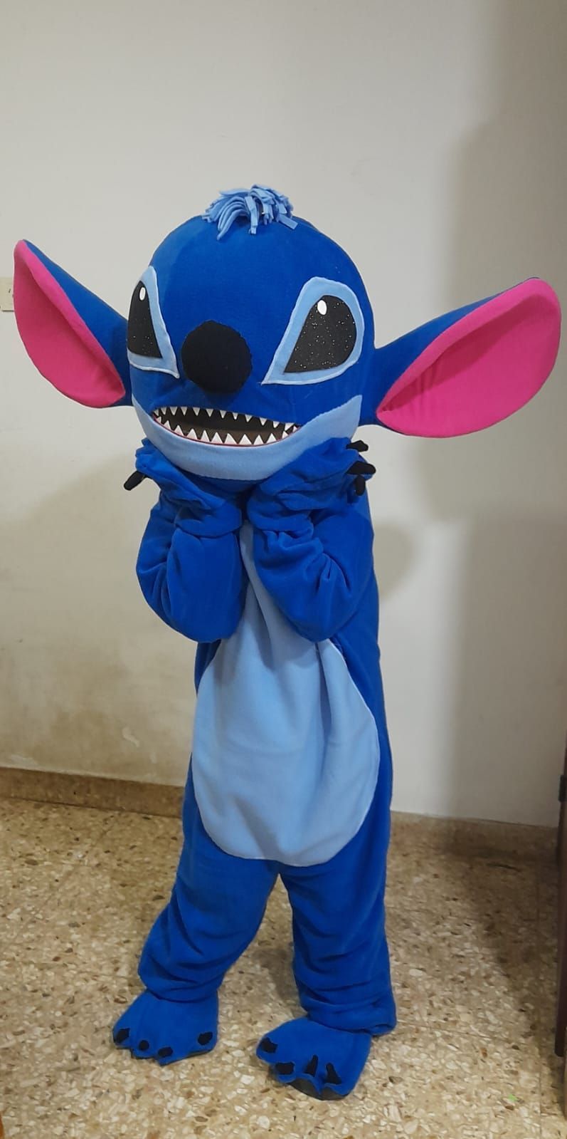 Una persona disfrazada de Stitch, de azul con orejas rosas, con las manos bajo la barbilla, en el interior de una vivienda.