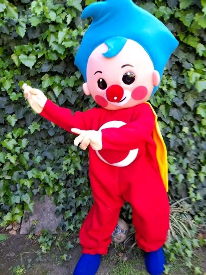 Mascota con mono rojo, pelo y zapatos azules, nariz y mejillas rojas, y capa amarilla. Posando con los brazos extendidos delante de la hiedra.