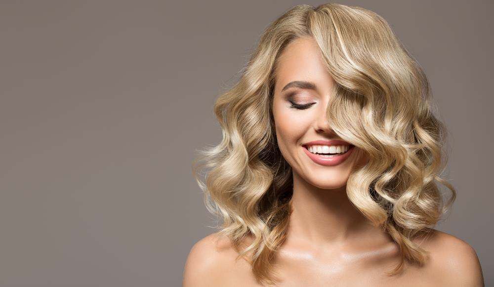 Smiling Blonde Woman with Voluminous Wavy Hair on A Gray Background — Fraser Shores Hair Design In Urraween, QLD