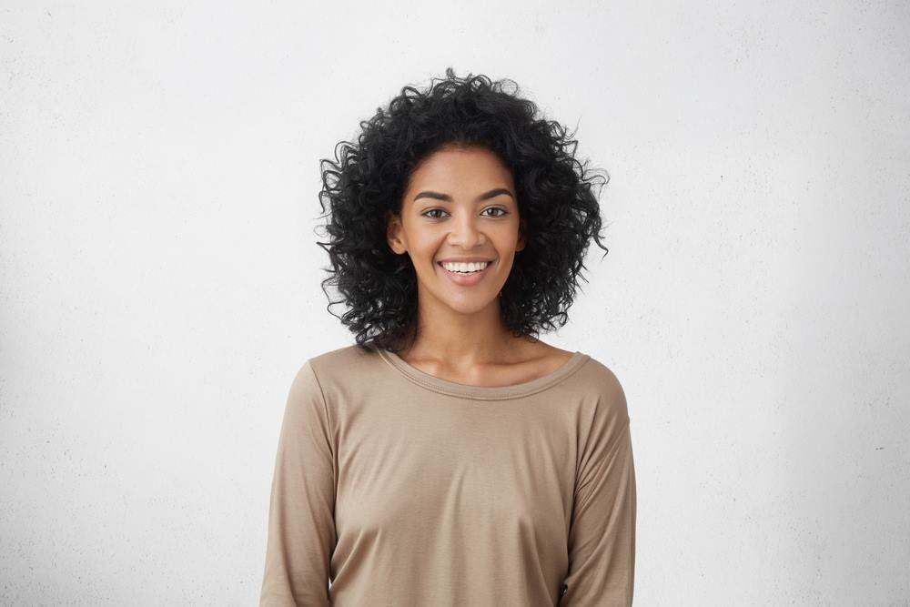 Smiling Person with Curly Hair Wearing a Beige Top Against a Light Background — Fraser Shores Hair Design in Urraween, QLD