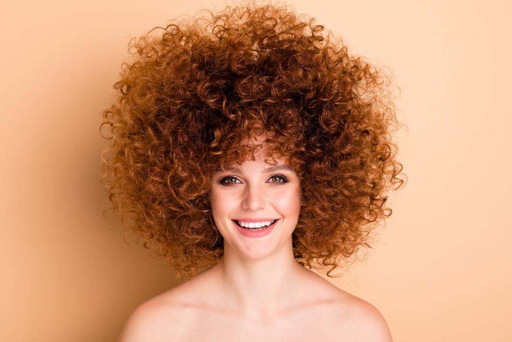 Smiling Person with Voluminous Curly Hair on A Warm Beige Background — Fraser Shores Hair Design in Urraween, QLD
