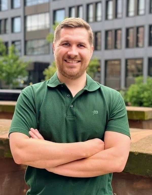 Man with crossed arms, wearing a green polo shirt, smiles outside a building.