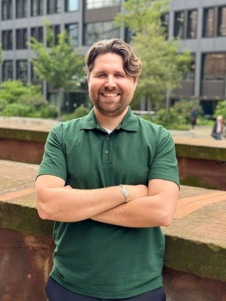 Man with crossed arms, wearing a green polo shirt, smiles outside a building.