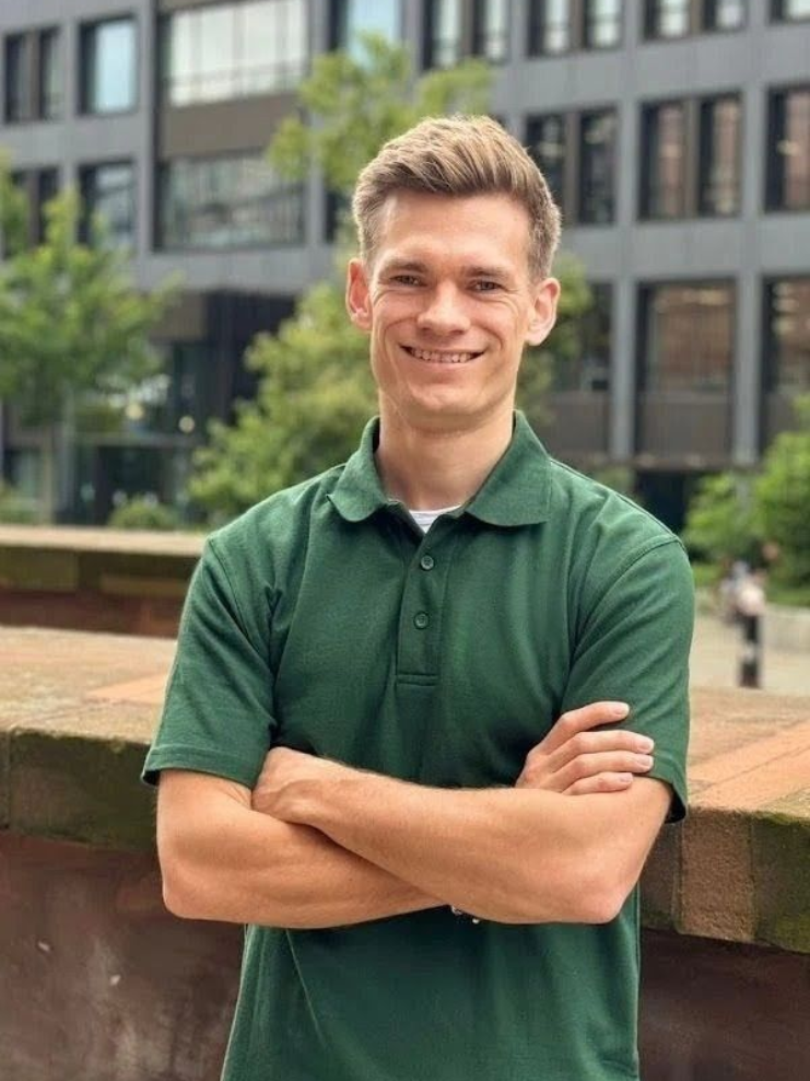 Man with crossed arms, wearing a green polo shirt, smiles outside a building.