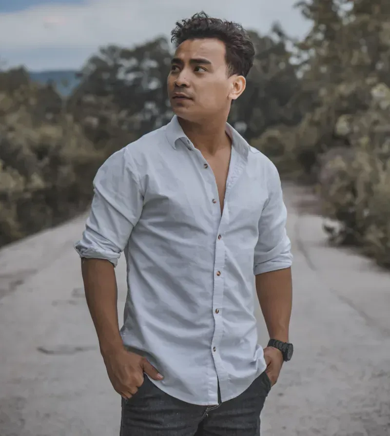 Man in a light blue shirt standing on a road, looking off to the side with trees and cloudy sky behind him