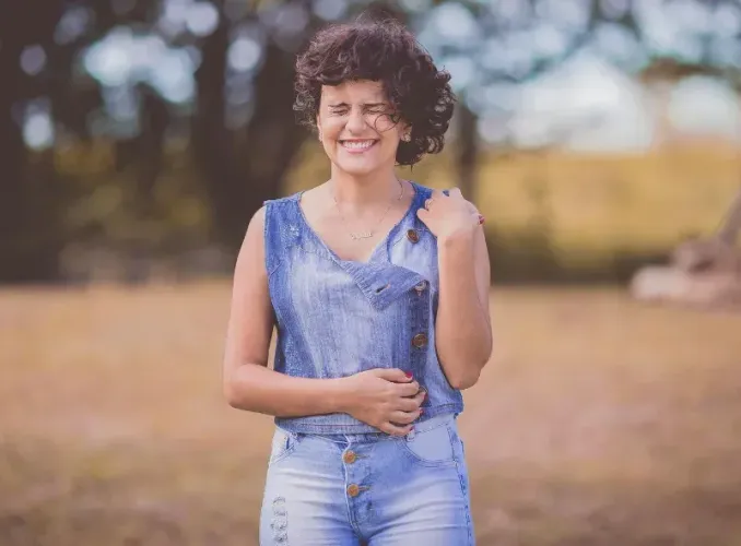 Smiling person in a denim outfit standing outdoors in a field with blurred trees in the background