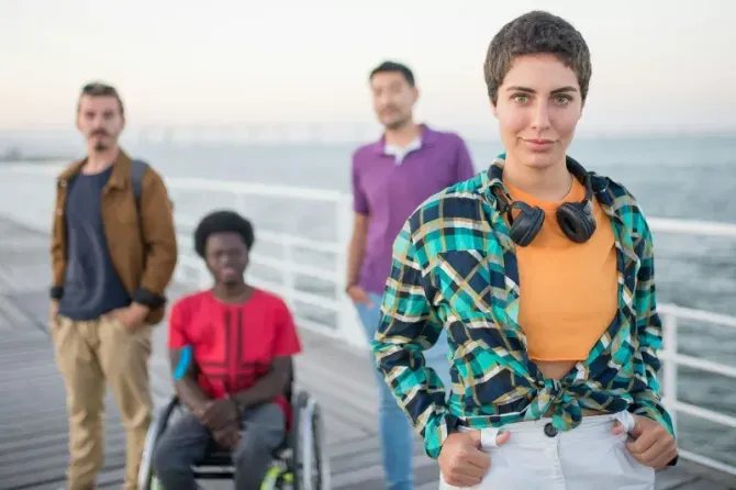 Four people on a waterfront boardwalk, with a woman in a plaid shirt in the foreground and city skyline behind.