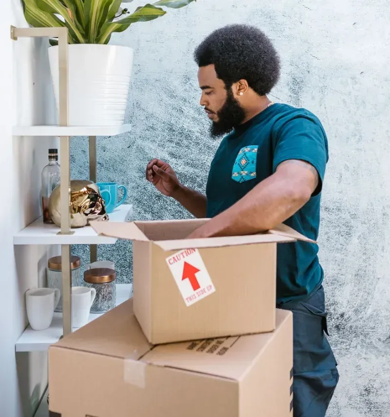 Man packing moving boxes beside a white shelf, placing items into an open box with a red up arrow label