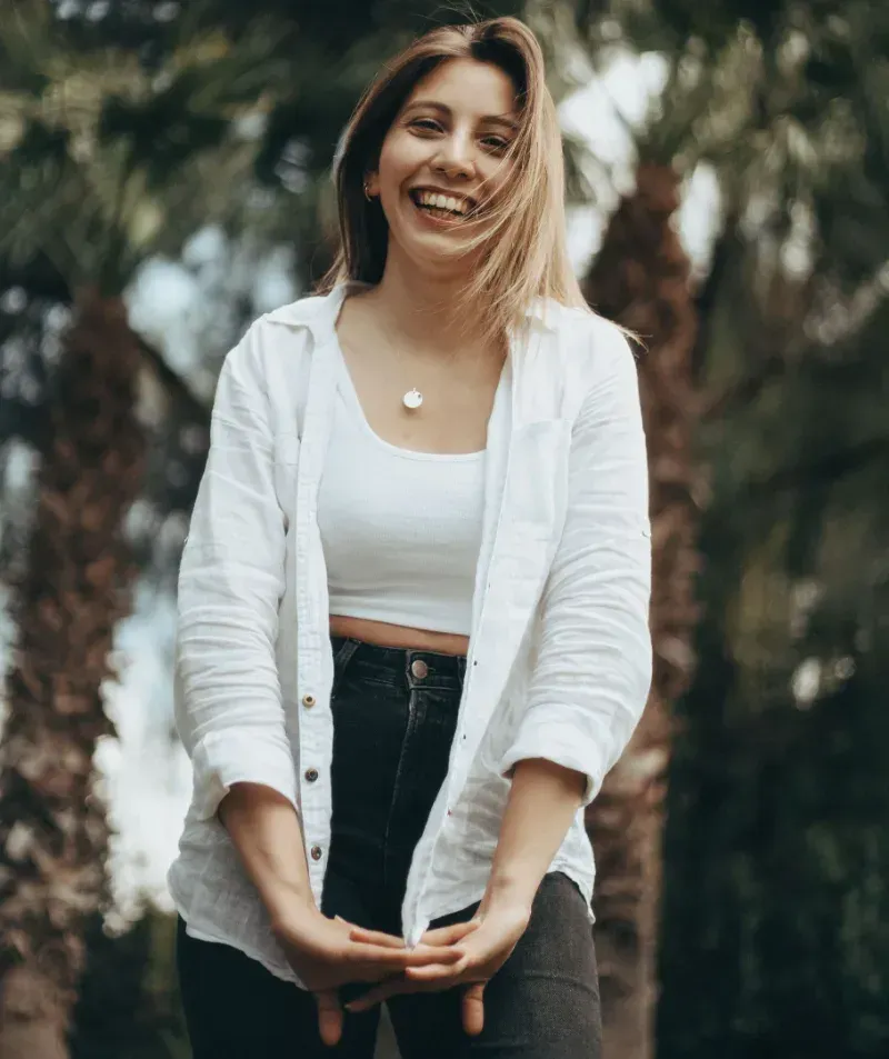Smiling woman in a white cardigan and black jeans standing outdoors with blurred palm trees behind her