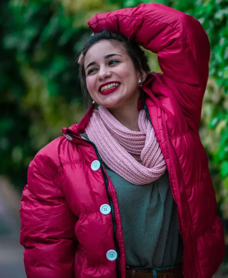 Smiling woman in a bright pink coat and scarf, posing outdoors with one arm raised