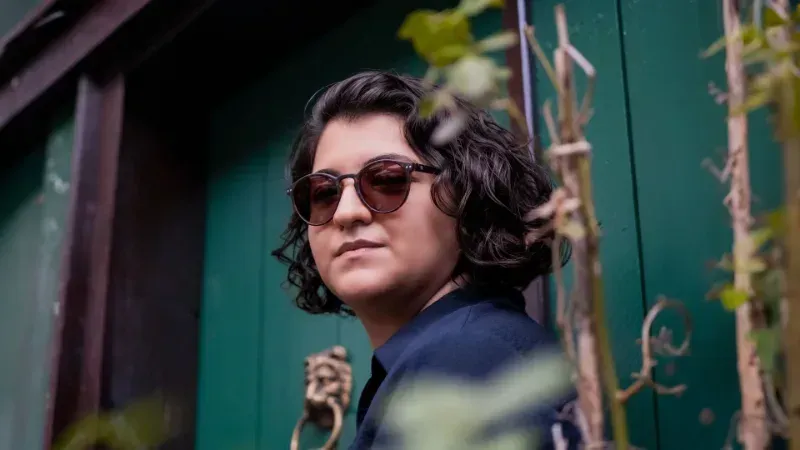 Person with curly dark hair and sunglasses stands by a green fence outdoors.