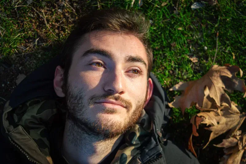 Person lying on grass in sunlight, looking upward, with dry leaves beside them.