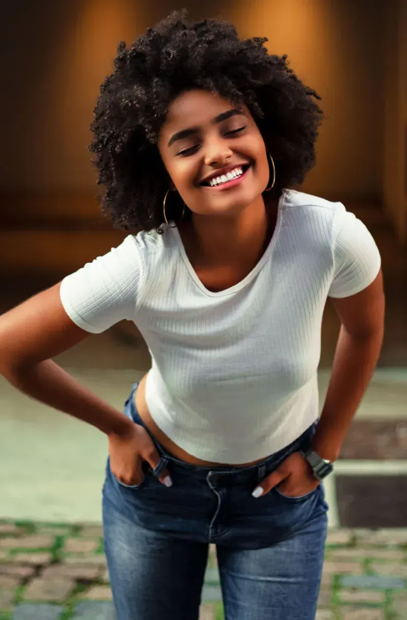 Smiling person in a white T-shirt and jeans leaning forward with hands on hips outdoors.