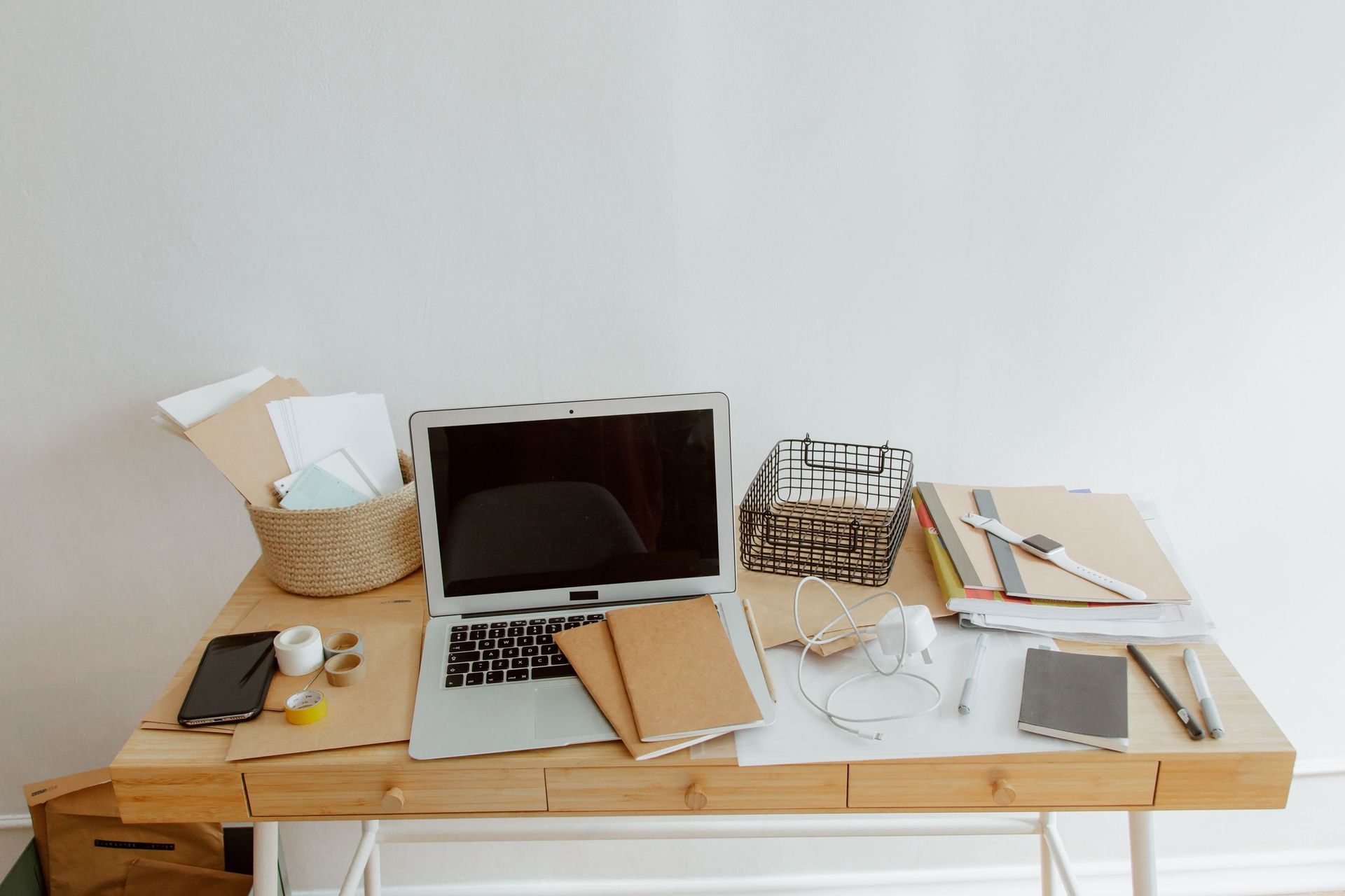 Messy wooden desk with open laptop, papers, phone, notebook, glasses, and a woven basket against a white wall