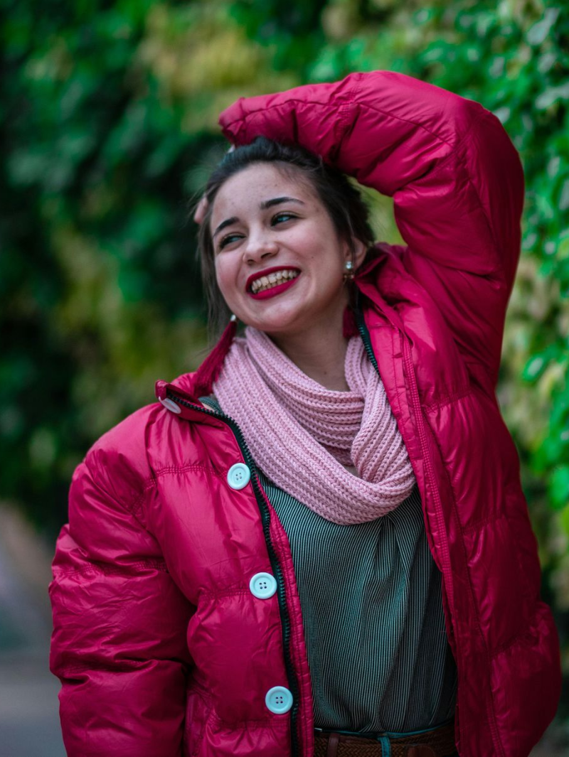 Smiling woman in a bright pink coat and scarf, posing outdoors with one arm raised