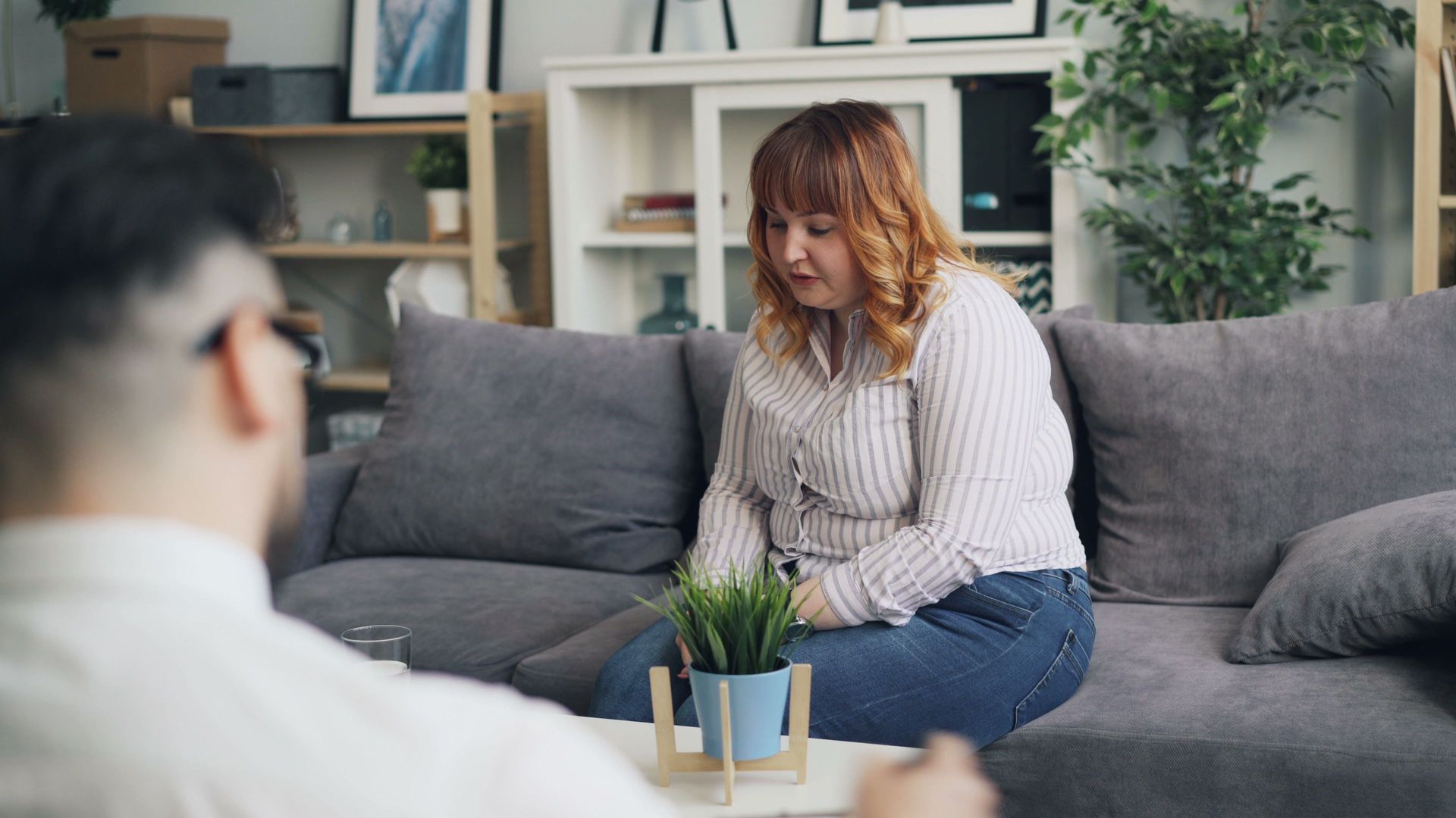 Woman sits on a gray couch in a living room, looking down with a pensive expression while another person is blurred in foreground.
