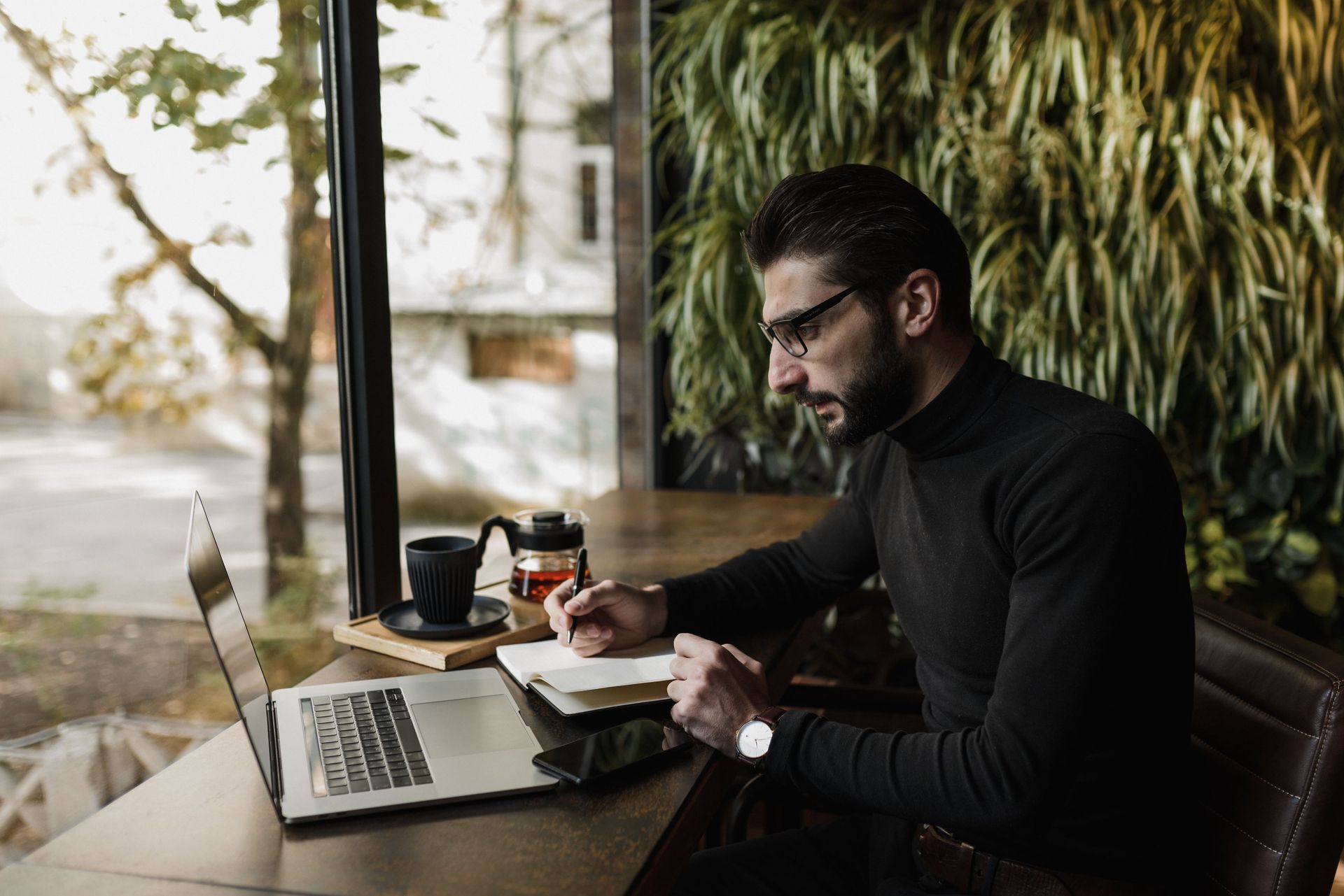Man in black sweater writing at a café table with a laptop, coffee, and notebook by a window
