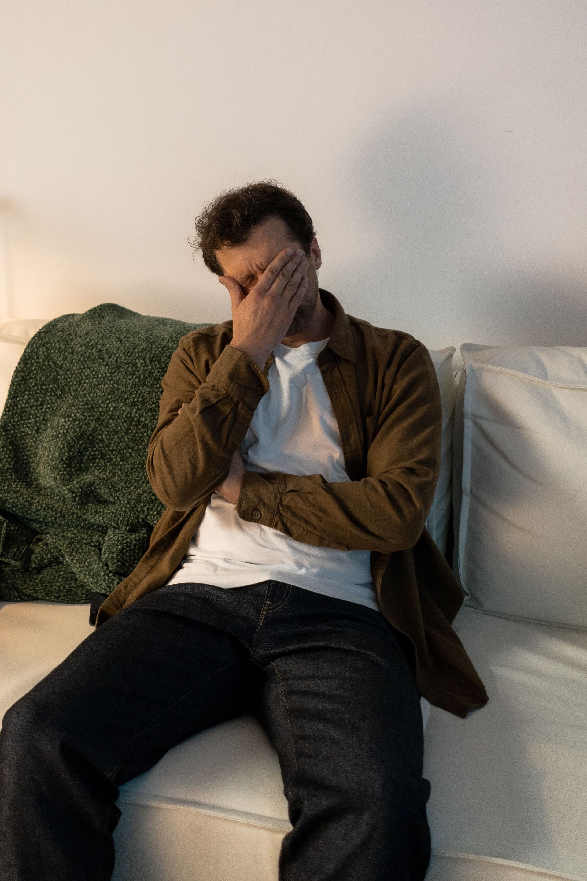 Person sitting on a white couch, covering their face with one hand, appearing distressed.