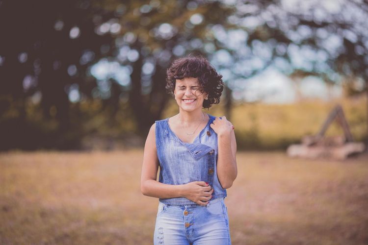 Smiling person in a denim outfit standing outdoors in a field with blurred trees in the background