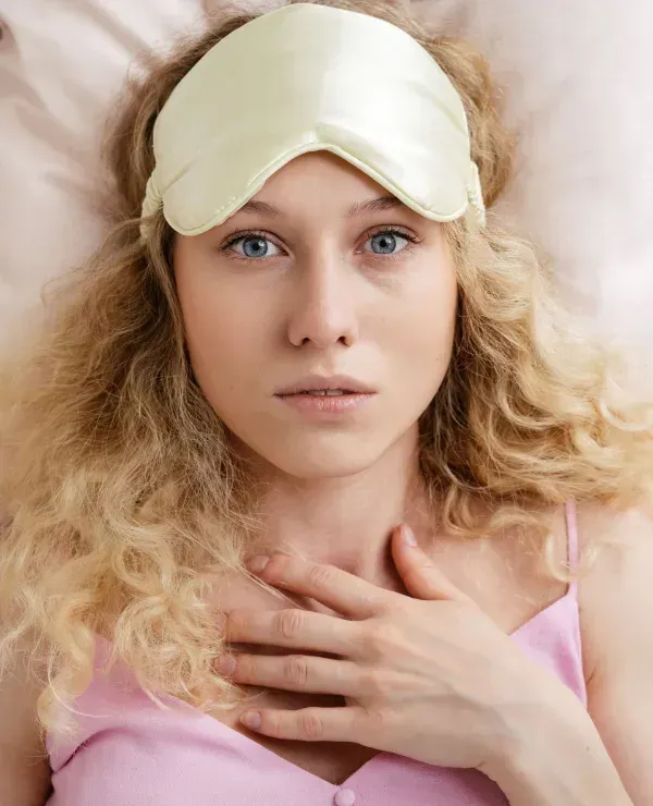 Person lying down with a sleep mask on their forehead and a hand on their chest, looking fatigued.