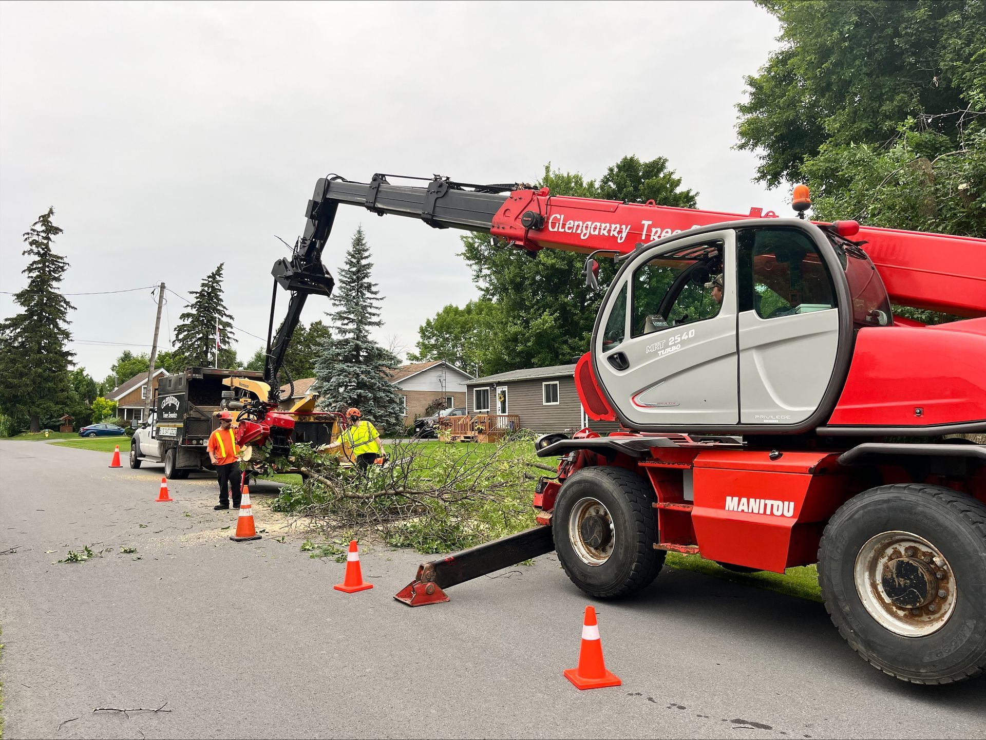 Red telehandler removes tree branches on a residential street. Workers in vests and a truck are present.