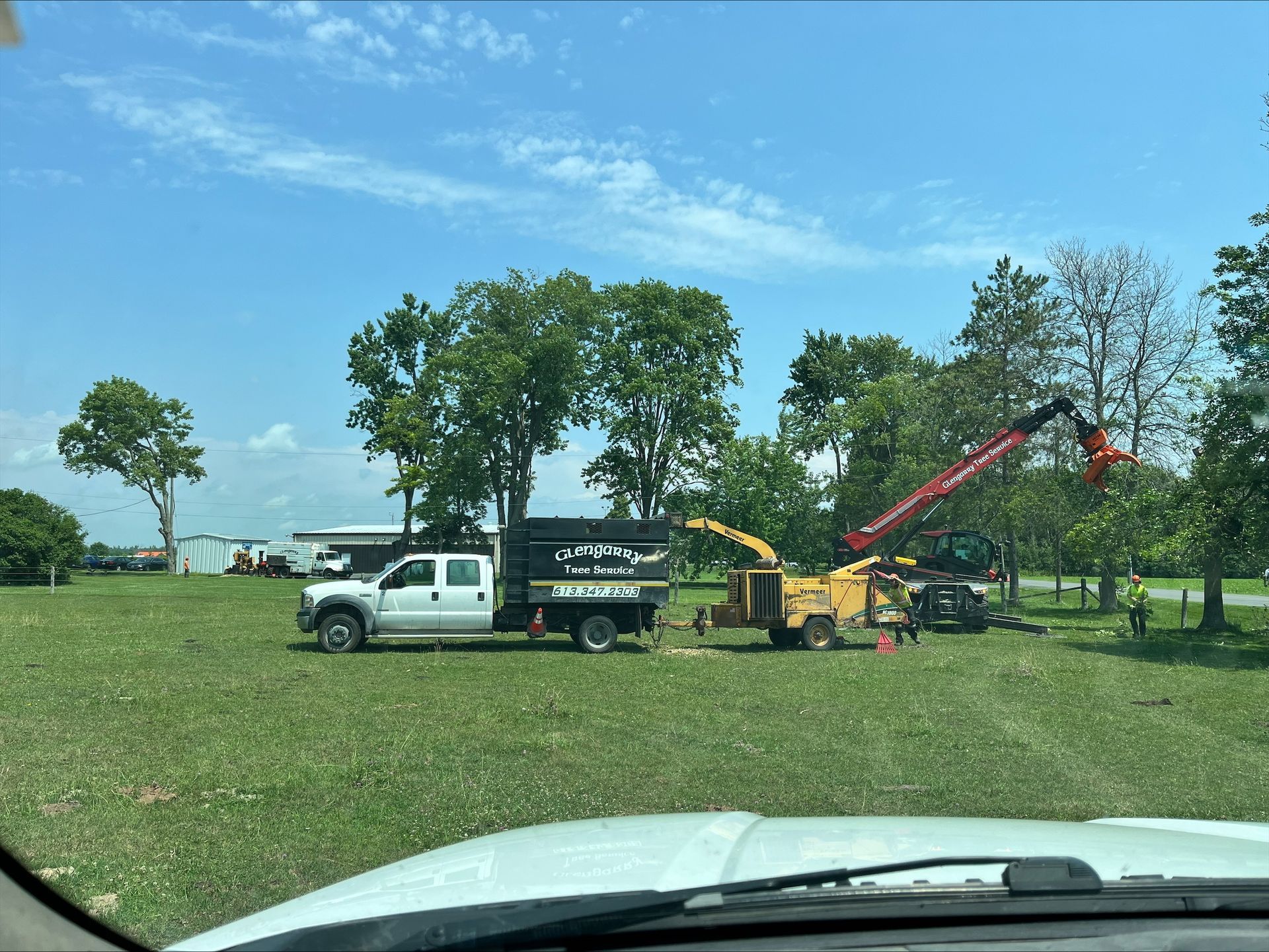 Tree service equipment in a grassy field on a sunny day. A truck, chipper, and crane are visible.