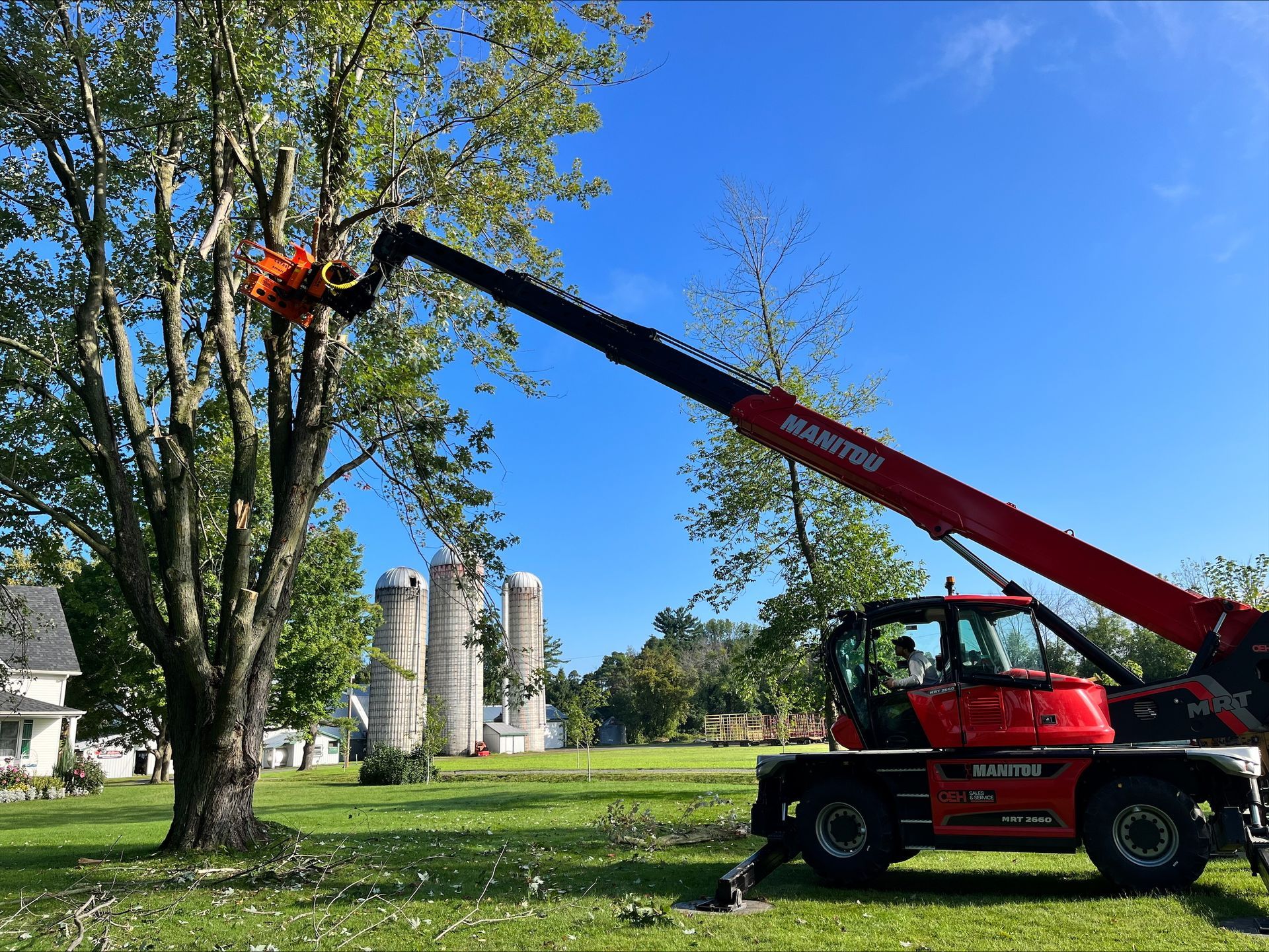 A red tree trimming machine cuts branches from a tree on a sunny day. Silos in the background.