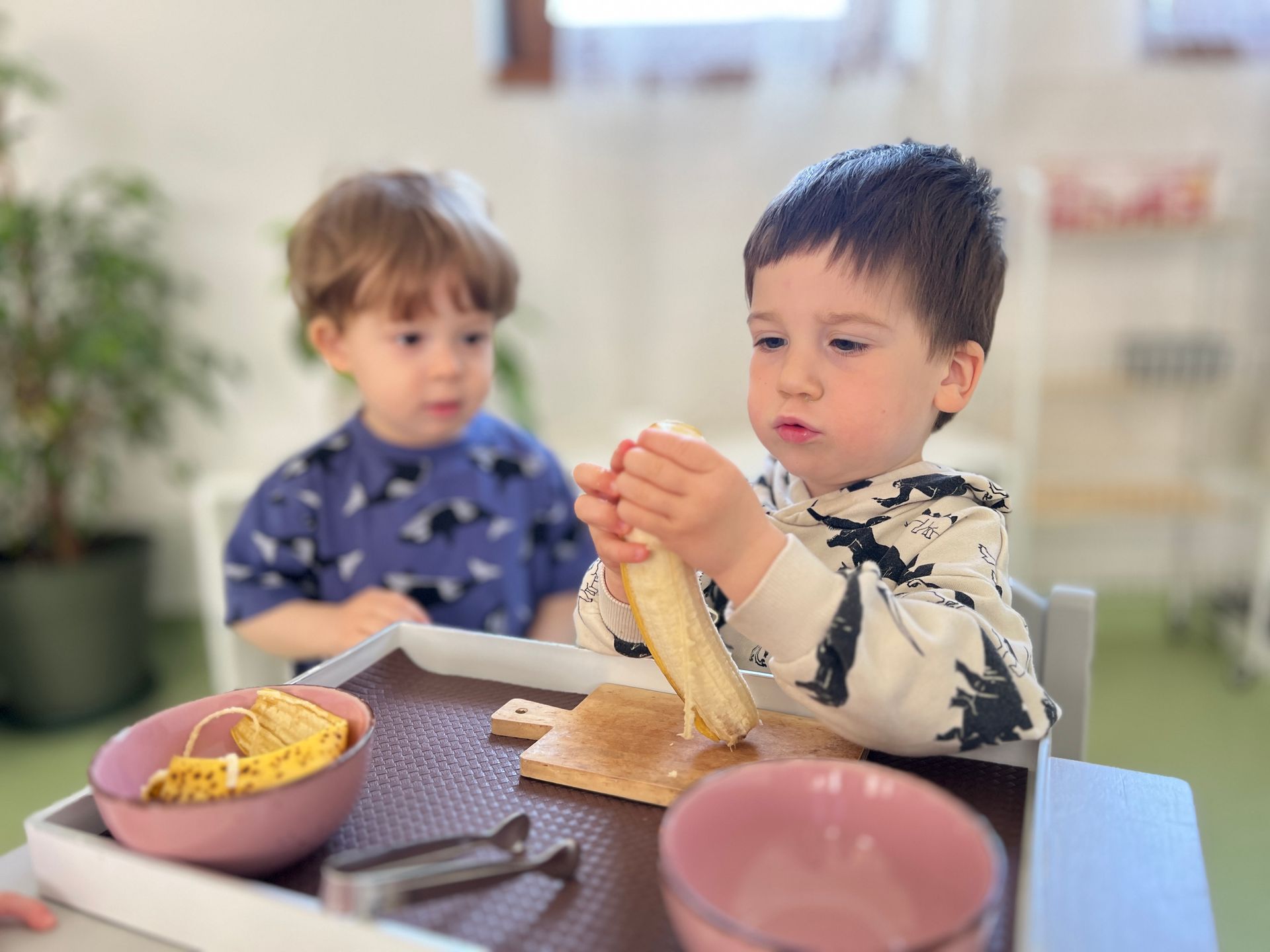 Two young boys are sitting at a table eating bananas.