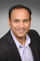 Smiling man, dark hair, wearing a blue button-down shirt and black blazer. Professional headshot.