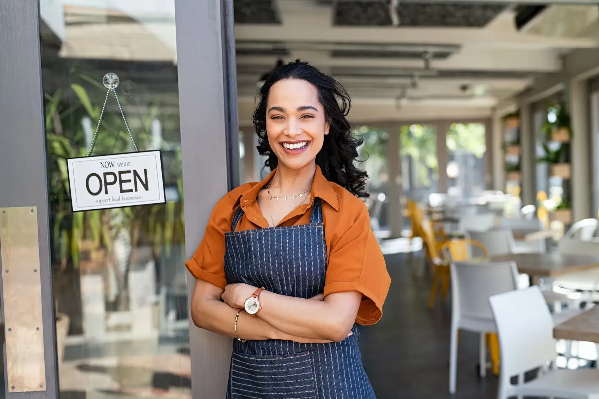 A woman in an apron is standing in front of an open restaurant door.