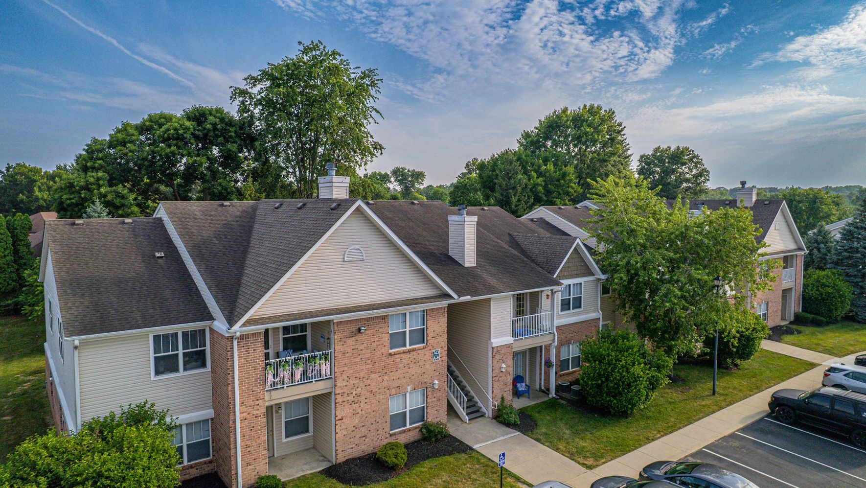 Apartment building with brick and siding, green trees, blue sky, and a parking lot with cars.