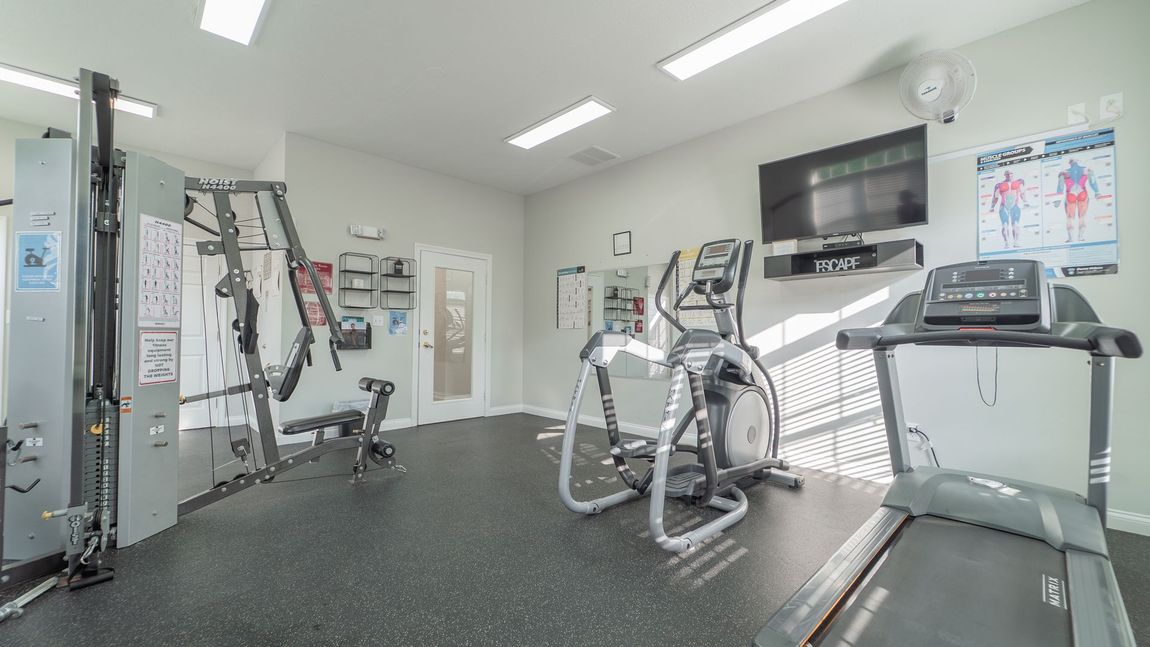 Gym with exercise equipment, including a treadmill, elliptical, and weight machines, against light walls.