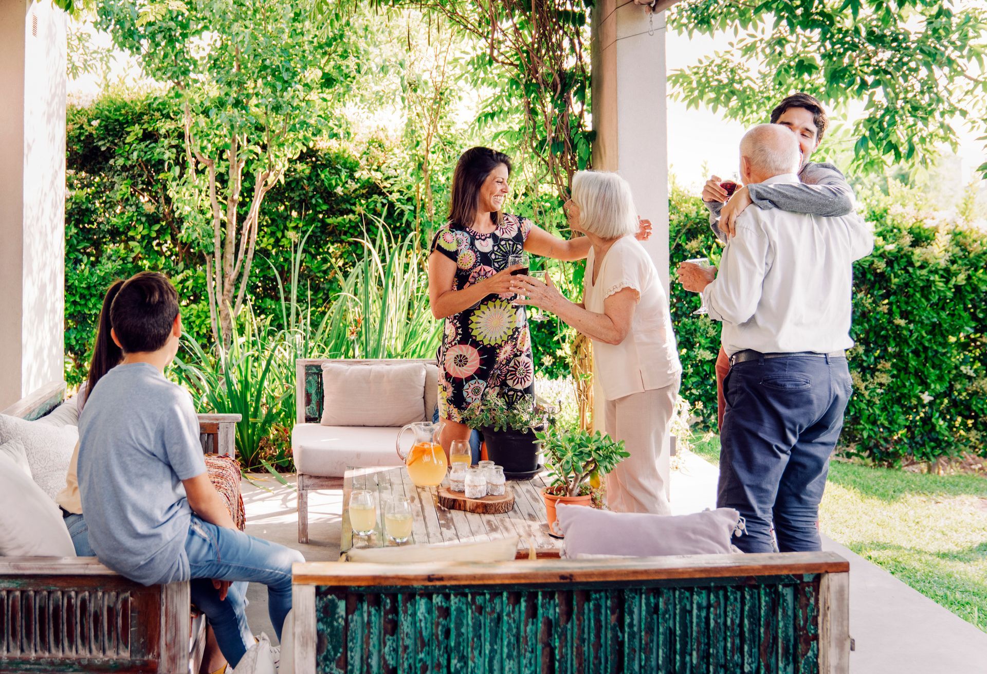 People chatting outdoors at a patio table with drinks and snacks, with greenery in the background
