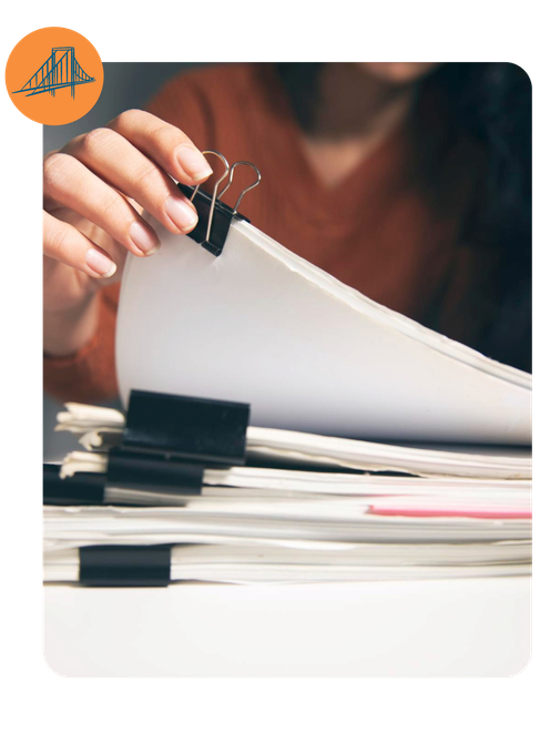 Hand holding a binder clip on stacked papers with a laptop in the background