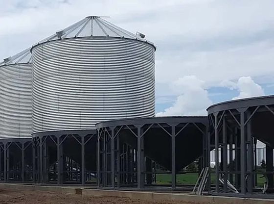 Large silver grain silo beside dark storage structures under a cloudy sky