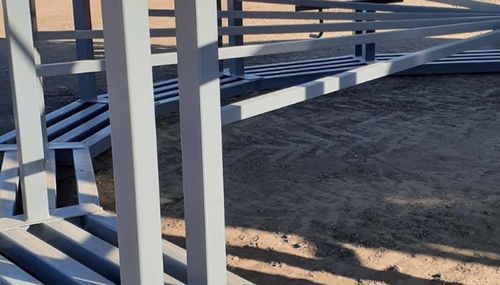 White bleachers with blue benches on a dirt field in bright sunlight