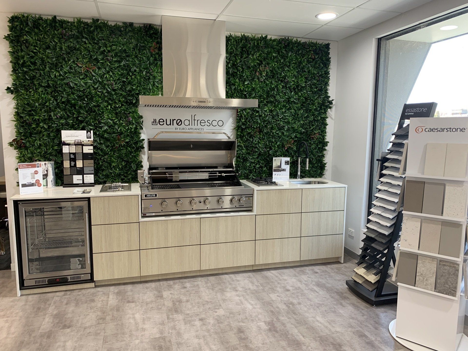 Outdoor kitchen display with grill, cabinets, and green wall backdrop.