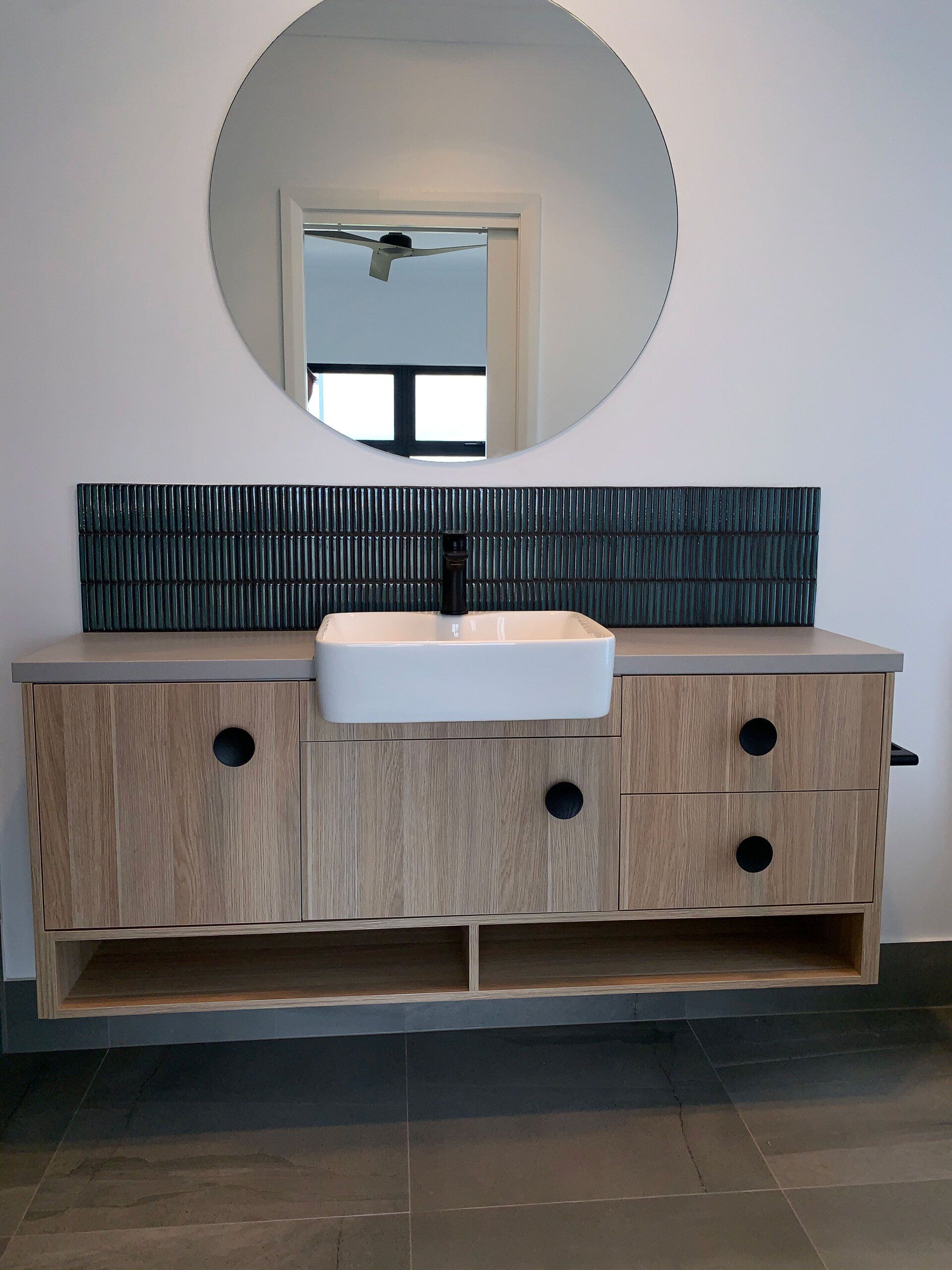 Floating wood vanity with white sink, black knobs, and textured tile backsplash, below a round mirror.