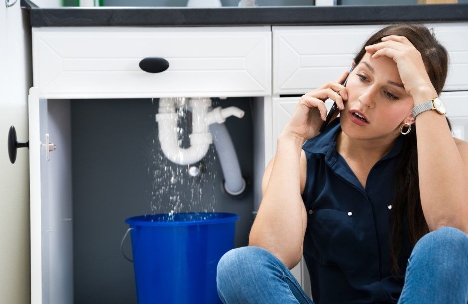 A woman is sitting under a sink talking on a cell phone.