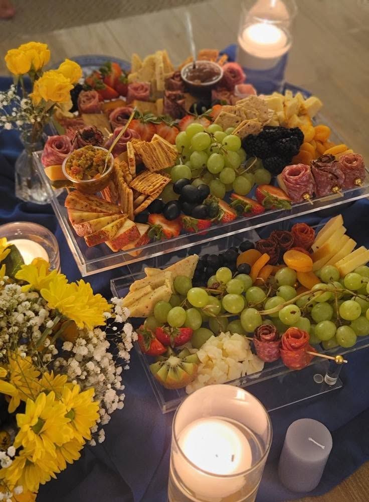 Candles lit beside a festive fruit and cheese platter with grapes, flowers, and crackers on a table