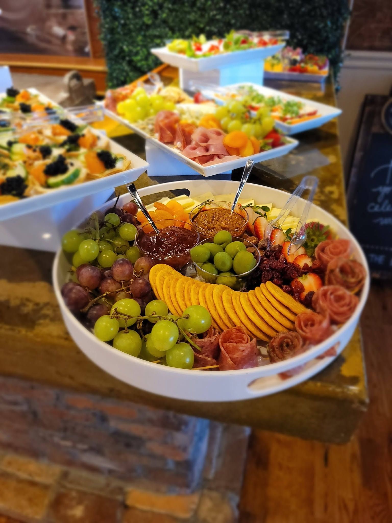 Bowls of colorful fruit and charcuterie on a wooden table at a buffet, including grapes, kiwi, and sliced salami
