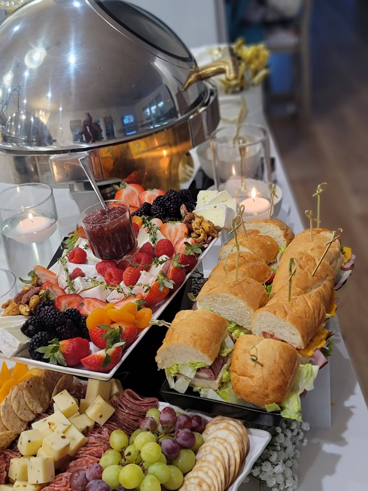 Buffet table with fruit, pastries, sandwiches, and cheese under a silver chafing dish lid