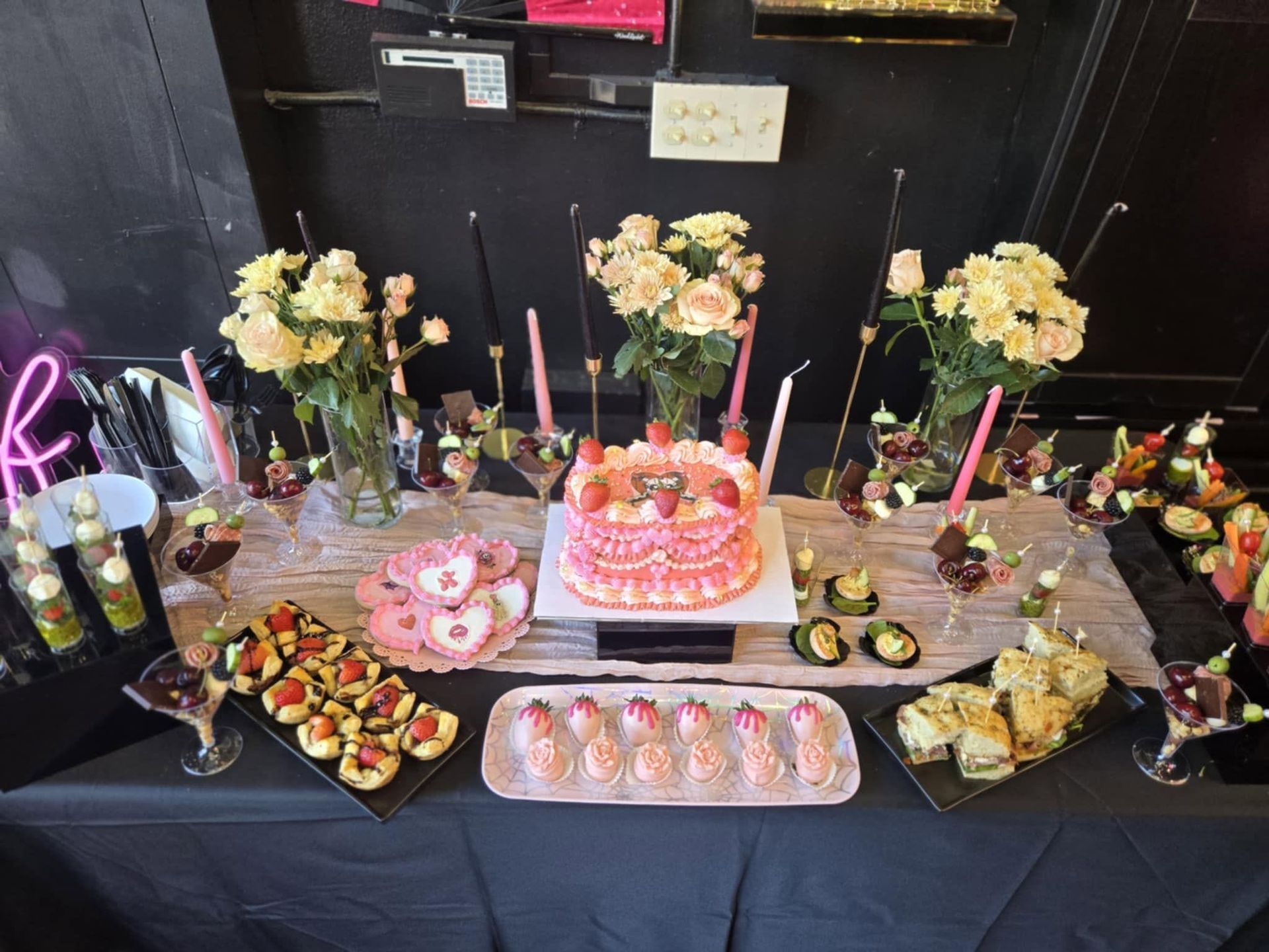 Dessert table with pink birthday cake, cupcakes, fruit platters, and flowers on a black backdrop