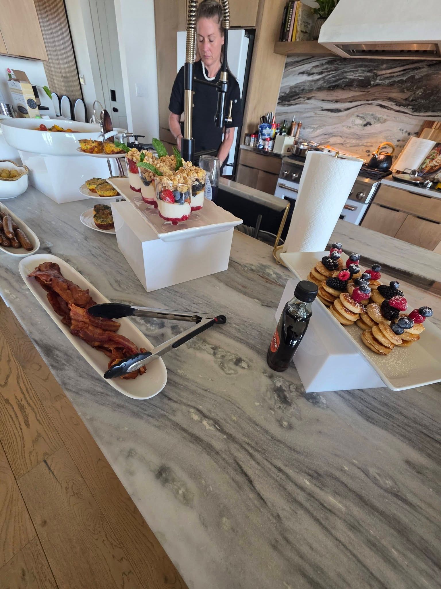Dessert buffet on a marble counter with cakes, pastries, fruit, and a man standing behind it.