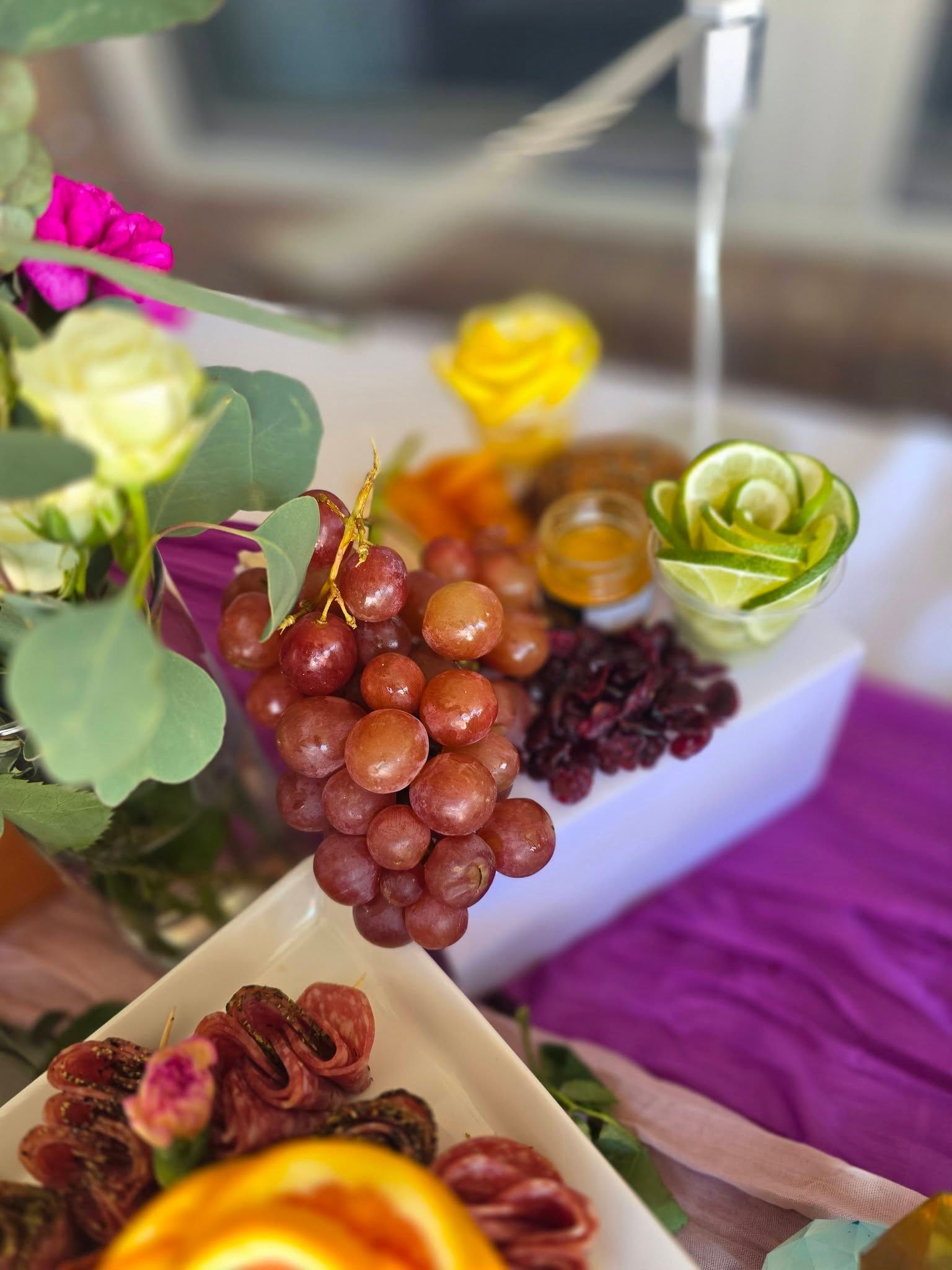 Fruit and flowers arranged on a table with purple cloth, including grapes, berries, and yellow blooms