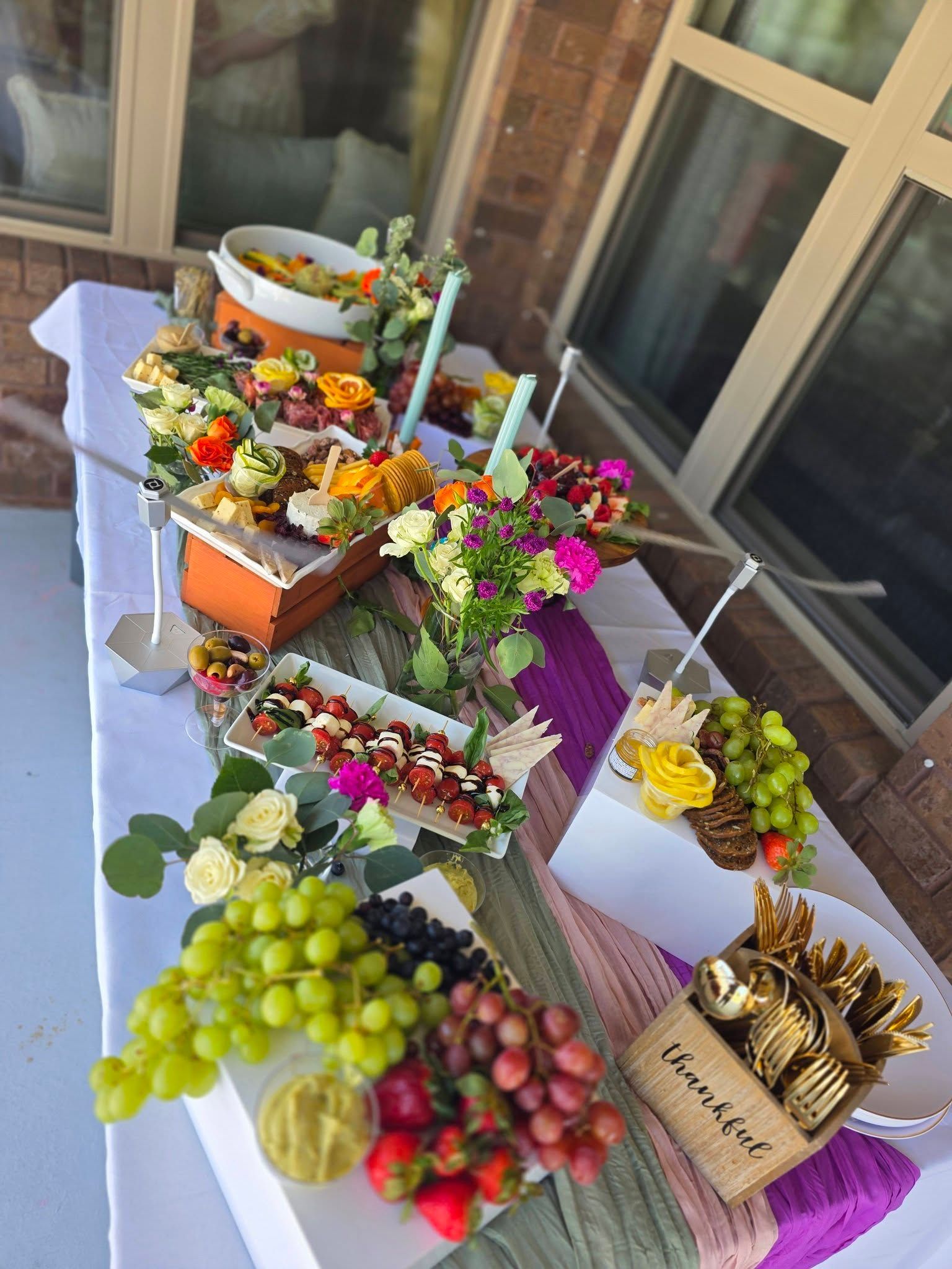 Outdoor table covered with colorful fruit, flowers, and decorative arrangements by a window