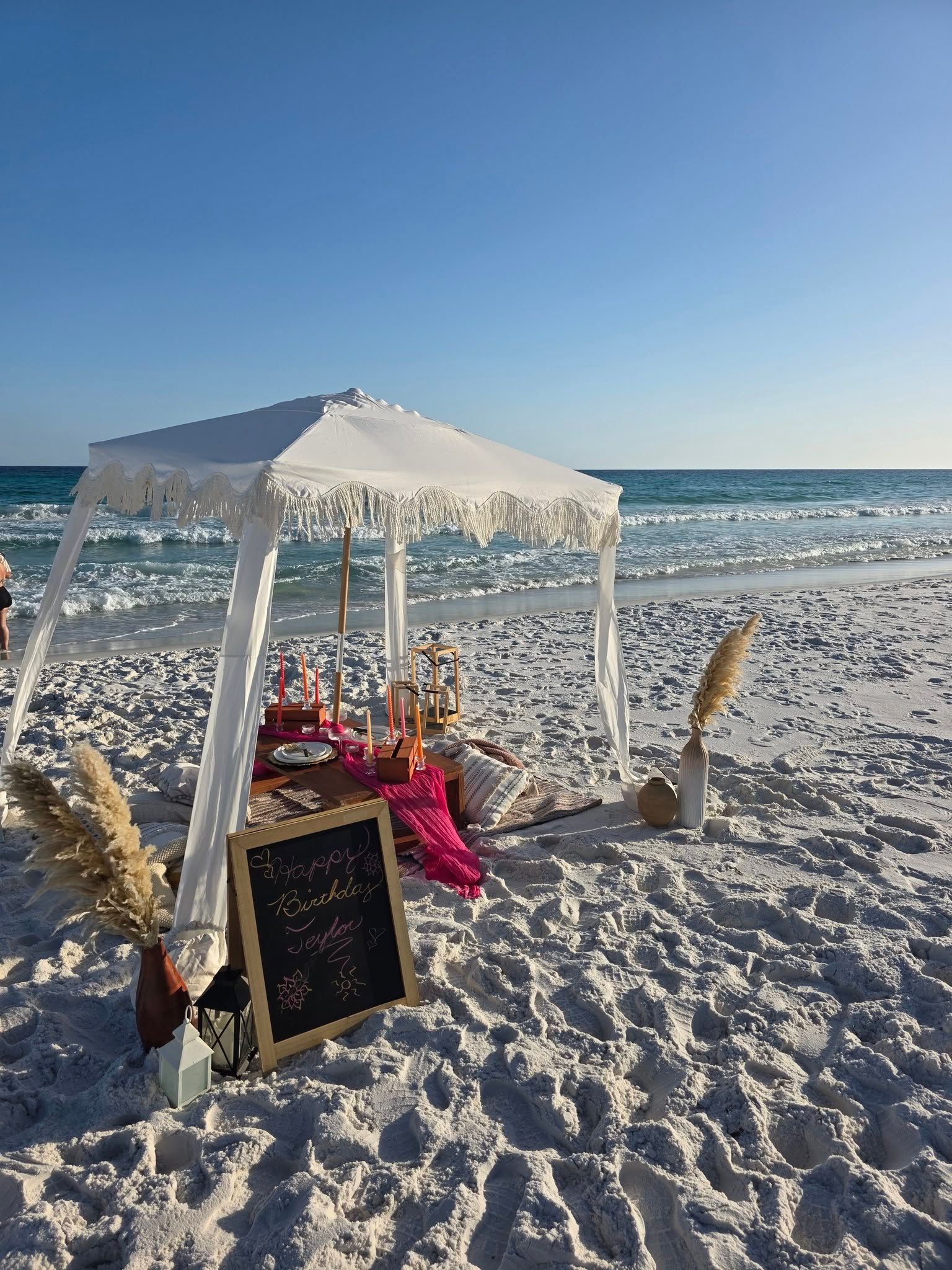 Beachside white canopy with pink blanket, flowers, and chalkboard sign on the sand by the ocean