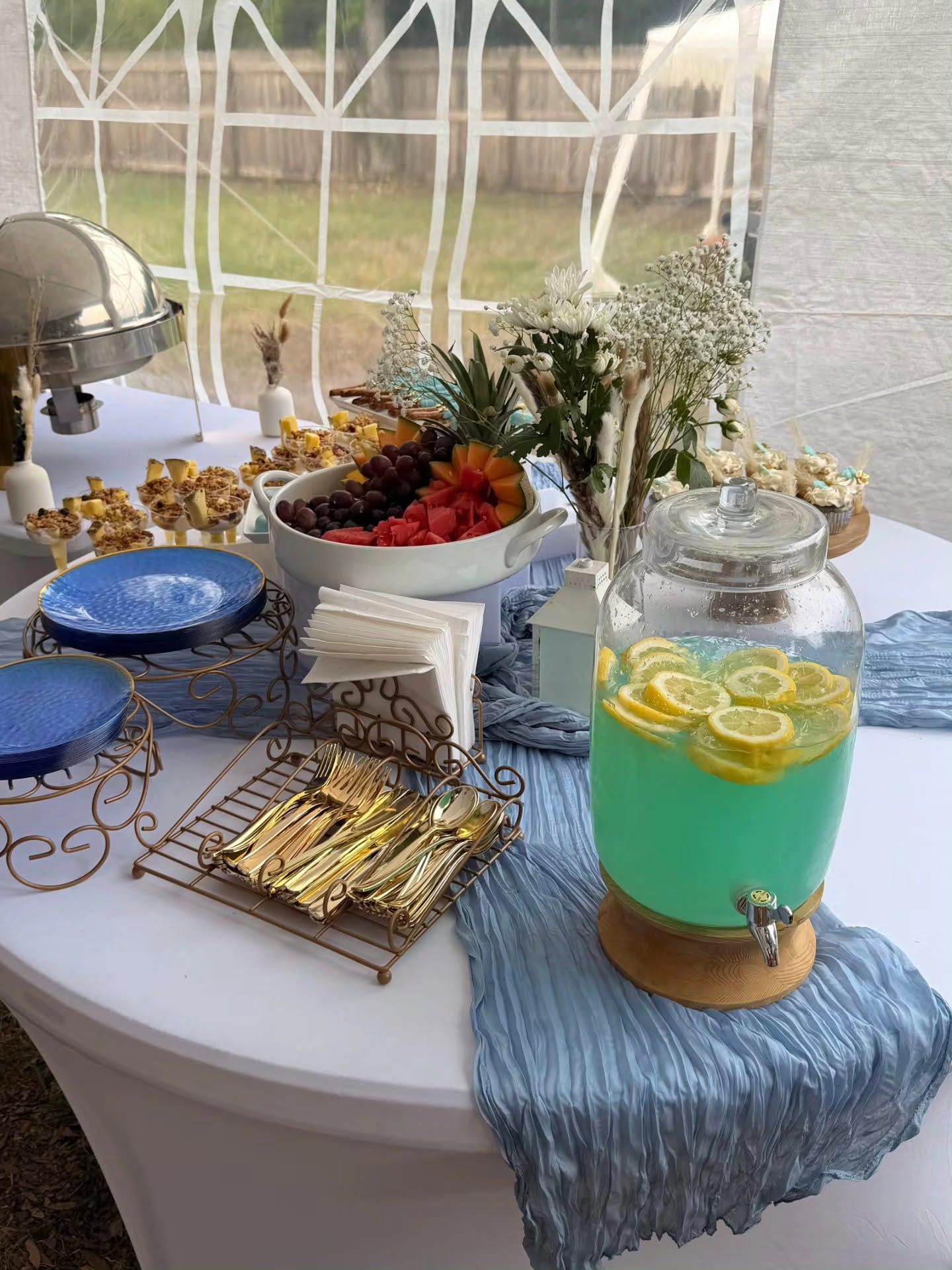 Dessert table with blue cake, fruit platter, cookies, and lemonade in a glass dispenser