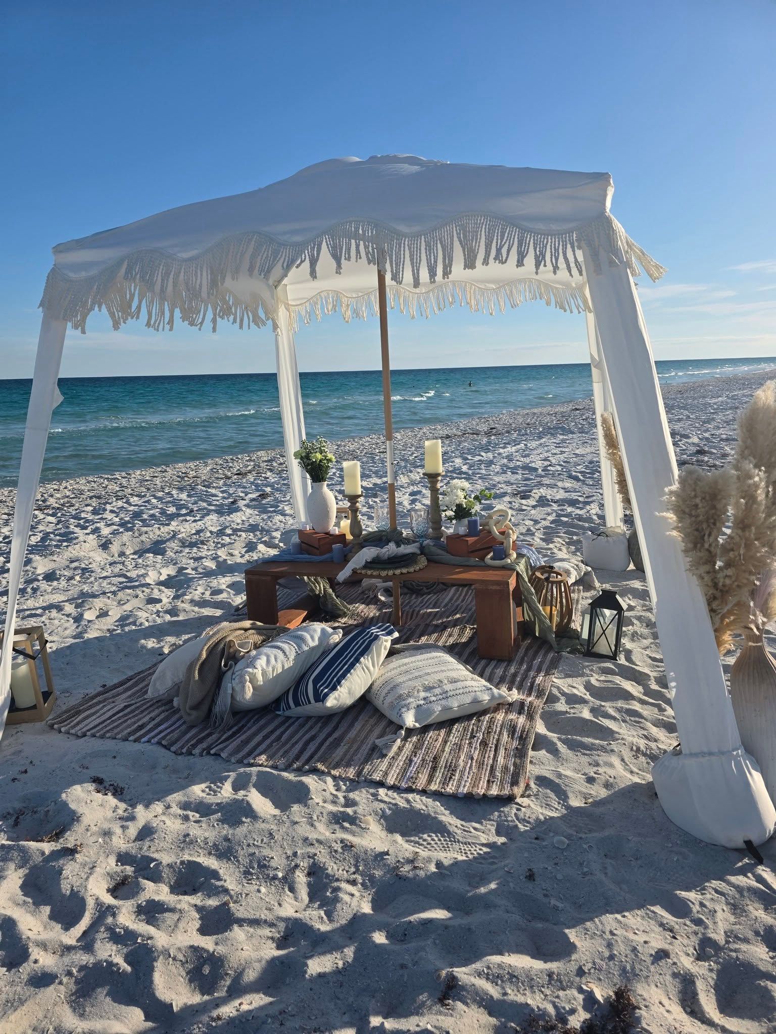 Beach canopy set up on a sunny sandy shore by the ocean, with cushions and table under a white drape.